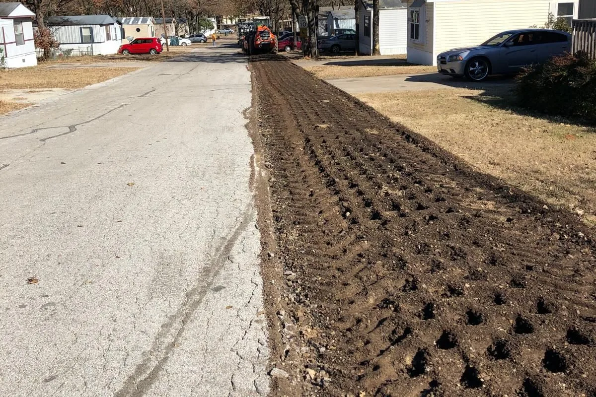 A residential street being rebuilt in place — half asphalt, half churned road base ready for LL-TECH integration. The materials you already own become the road.